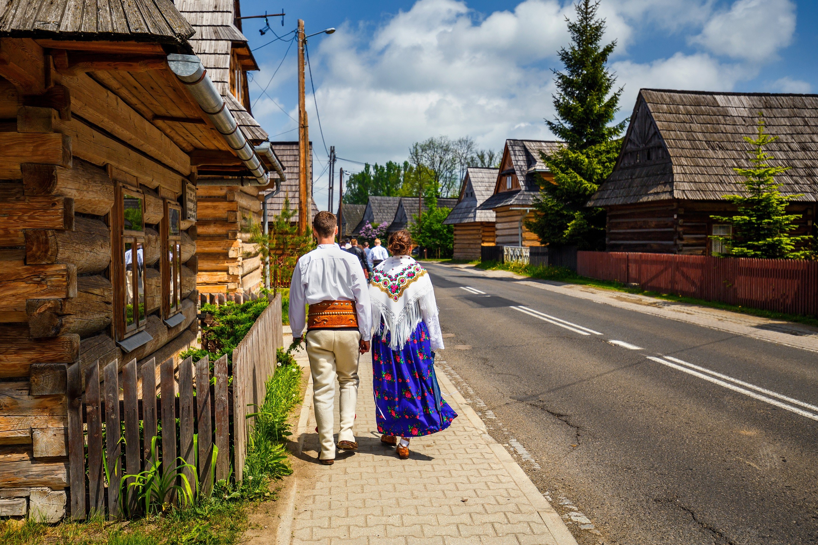 Para ubrana w regionalny strój góralski idzie wzdłuż ulicy w Chochołowie. Do ulicy przystają ściany drewnianych chałup.